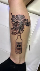 Черно-белая татуировка на голени: бутылка Tito’s Handmade Vodka с букетом цветов, выходящих из горлышка — реалистичный стиль с элементами ботанической графики