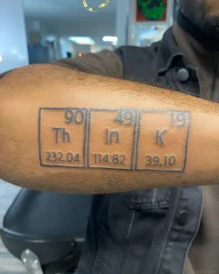 Черно-серая татуировка на предплечье в виде трёх ячеек периодической таблицы Менделеева — торий (Th), индий (In), калий (K) — с атомными номерами и массами, выполненная в стиле минимализма с чёткими контурами
