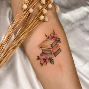 Нежная татуировка с изображением стопки книг и розовых цветов на предплечье, выполненная в стиле акварельной миниатюры