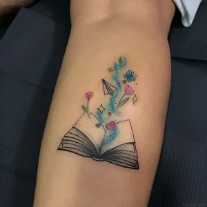 Яркая татуировка на предплечье с открытой книгой, из которой вырывается поток фантазии: цветы, бабочки и бумажный самолетик