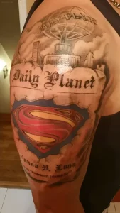 Детальная татуировка на плече мужчины: здание Daily Planet над разорванной тканью, под которой виден цветной щит Супермена, внизу имя и надпись 'You are the reason I could fly'