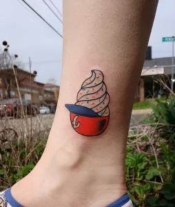 татуировка с рисунком мягкого мороженого со sprinkles в красной чашке на ноге человека, фотография для статьи о тату ко Всемирному дню мороженого