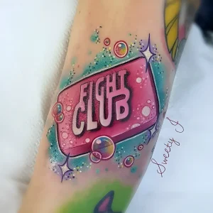 Татуировка на предплечье с яркой надписью FLIGHT CLUB, окруженная мыльными пузырями, блестками и звездочками в стиле нео-пастелли