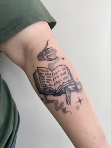 Черно-белая татуировка на руке с изображением открытой книги, из которой исходит дым, и звезд, с текстом на английском языке о преодолении трудностей