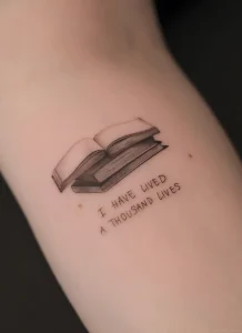 Черно-белая татуировка на руке с изображением раскрытой книги и текстом I HAVE LIVED A THOUSAND LIVES, посвященная Общероссийскому дню библиотек 27 мая