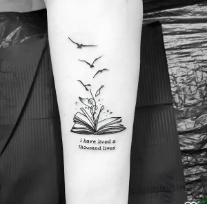 Черно-белая татуировка на предплечье с изображением книги, из которой вылетают птицы, и надписью I have lived a thousand lives