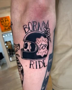 Черно-белая татуировка в стиле American Traditional на руке: байкерский шлем, надпись Born to Ride, цветок