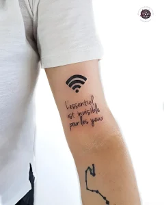 Черно-белая татуировка на предплечье: сверху — стилизованный значок Wi-Fi, под ним — рукописная надпись на французском языке «L'essentiel est invisible pour les yeux», ниже — контурный силуэт сердца. Минималистичный стиль, тонкие линии, чёткая