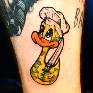 Яркая татуировка на ноге в стиле нео-традишнл: желтая утка в белом колпаке с ножами, зелеными акцентами и надписью PUNK