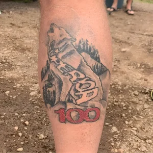 Крупный план татуировки на бедре: рыкающий волк с цифрами 100 и стилизованным лесом, выполненная в черно-белой гамме с красными акцентами