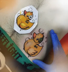 Свежевыполненная татуировка с желтой уточкой в очках-гoggles и надписью 'Duck' на коже рядом с трафаретом