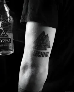 Черно-белое фото татуировки на предплечье с надписью Techno и геометрическим узором, рядом бутылка водки