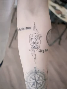 Татуировка с масками комедии и трагедии, компасом и надписью 'smile now cry later' на предплечье