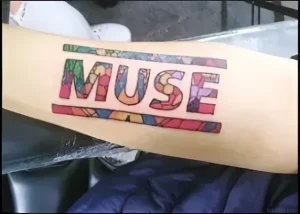 Яркая татуировка с надписью MUSE, выполненная в стиле цветного витража с разноцветными фрагментами на предплечье