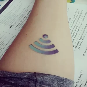 Крупный план цветной татуировки в виде значка Wi-Fi на предплечье человека, лежащего на учебном материале. Тату выполнена в градиенте от фиолетового до бирюзового, с чёткими контурами и современным минималистичным дизайном.