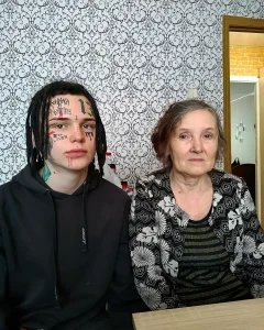 Портрет молодого человека с черными дредами и лицом, полностью покрытым текстовыми татуировками и символами, сидящего рядом со своей бабушкой на фоне цветочного обоев