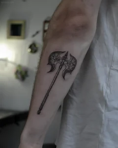 Татуировка топора в стиле blackwork на предплечье мужчины, выполненная с использованием штриховки и узоров на лезвии