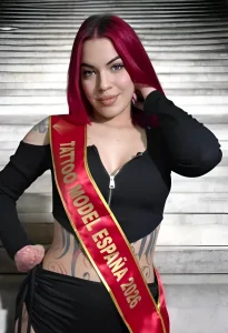 Девушка с ярко-бордовыми волосами и саше Tattoo Model España 2026 в черном облегающем костюме демонстрирует масштабную татуировку в стиле японского ирэдзуми на животе и боках. На фоне металлической стены она уверенно позирует, подчеркивая эстетику
