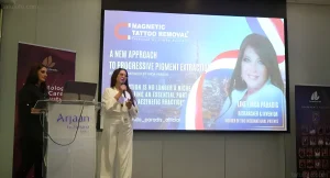 Изобретательница магнитного удаления тату Линда Парадис выступает с презентацией на сцене конференции по дерматологии и бьюти в Дубае, на экране за ней — слайд с описанием технологии и её портретом