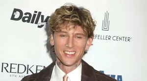 Портрет рэпера Machine Gun Kelly с блонд-волосами и пирсингом на фоне логотипа Daily Front Row, на шее видны черные татуировки