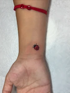 Крупный план женской руки с красным шнурком, на запястье которой выполнена яркая цветная татуировка божьей коровки в стиле микро-реализм