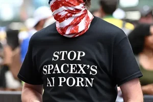 Человек в чёрной футболке с белым текстом 'STOP SPACEX’S AI PORN' и красно-белой повязкой на лице, участвующий в протесте в Нью-Йорке