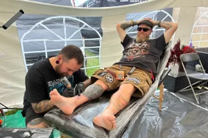 Тату-мастер наносит татуировку на голень посетителя фестиваля Download Festival 2026 в палатке
