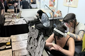Тату-мастер наносит татуировку клиенту, который сидит в VR-очках на Leeds International Tattoo Expo 2026. На переднем плане — стенд с эскизами, на заднем — другие мастера и клиенты.