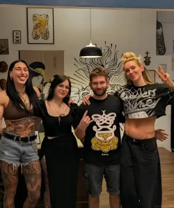 Фотография группы людей, включая Rhea Ripley и Charlotte Flair, в студии тату-мастера. Rhea Ripley показывает татуировку на животе.
