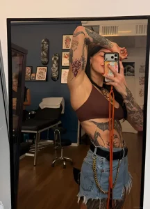 Rhea Ripley taking a mirror selfie in a tattoo studio in Italy, showing off her extensive sleeve and torso ink while wearing a brown top and denim skirt.