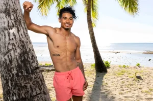 Участник шоу Love Island USA Синсер Риа стоит у пальмы в розовых шортах, на его правом боку видна татуировка с римскими цифрами II-XXXIV