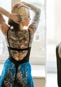 Фотография тату-модели Суки (Suki) в полный рост, демонстрирующая масштабную татуировку на спине и руках в стиле реализм. Модель одета в откровенный черный корсет и синие кружевные легинсы, стоит спиной к камере у окна.