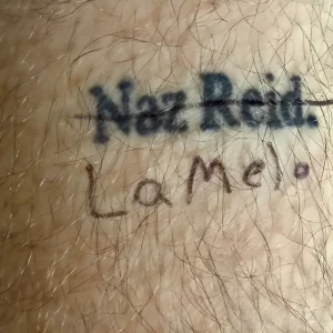 Крупный план татуировки на волосатой коже: имя 'Naz Reid' выполнено жирным шрифтом, под ним — надпись 'LaMel' тонкими линиями. Чёрные чернила, реалистичный стиль, видны волосы и текстура кожи.