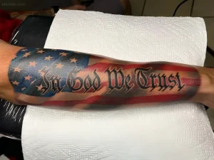 Свежая цветная татуировка на предплечье: надпись In God We Trust в готическом шрифте на фоне развевающегося флага США
