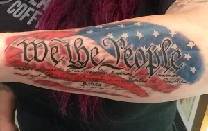 Крупный план татуировки на предплечье с текстом "We the People" над флагом США, выполненной в стиле акварель с использованием черного шрифта