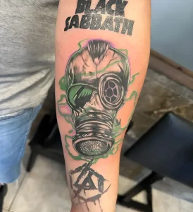 Детальная татуировка на руке с изображением противогаза, надписи Black Sabbath и пентаграммы, выполненная в стиле нео-традишнл с яркими акцентами