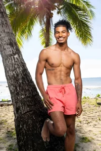Синсер Риа из реалити-шоу Love Island USA стоит у пальмы на пляже в розовых шортах, демонстрируя татуировку с римскими цифрами на правом боку