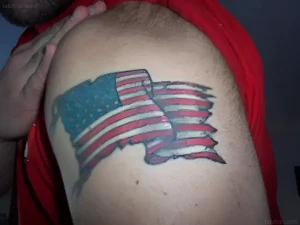 Крупный план татуировки на плече мужчины: американский флаг в стиле American Traditional, изображенный в виде разорванного полотнища, символизирующего патриотизм и свободу.