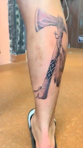 Крупный план татуировки с изображением боевого топора с перьями и орнаментом на голени мужчины