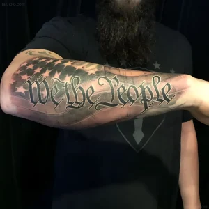 Мужская татуировка с текстом 'We the People' в готическом шрифте на фоне стилизованного флага США, выполненная в технике реализм с акварельными разводами