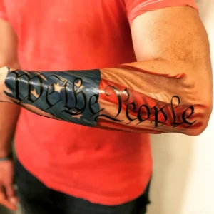 Мужская татуировка с текстом We the People на фоне американского флага на предплечье