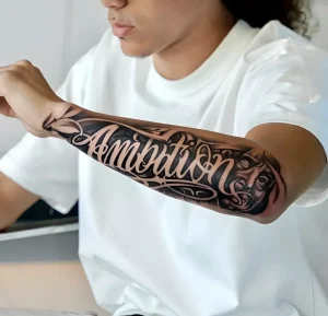 Крупный план предплечья с объемной татуировкой слова Ambition, выполненной в стиле нео-традишнл с элементами граффити и 3D-эффектом
