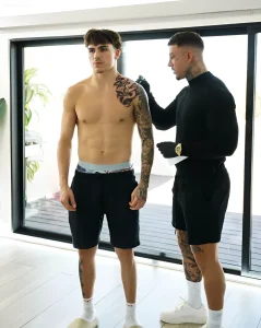Фотография тату-мастера Vendler Tattoo в черной одежде и перчатках, работающего с клиентом в студии. Клиент стоит без рубашки, демонстрируя татуировку на плече и руке.