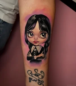 Детальная цветная татуировка Wednesday Addams с большими глазами и косичками в стиле Neo-Traditional на руке, выполненная мастером с подписью внизу