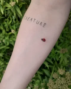 Фотография татуировки на женском предплечье: надпись Creature тонким шрифтом и маленькая красная божья коровка на фоне зелени