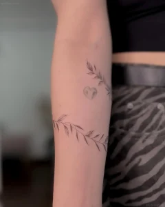 Черно-белая татуировка в стиле минимализм на внутренней стороне предплечья: тонкие ветки с листьями образуют браслет, в центре — стилизованное сердце с негативным пространством
