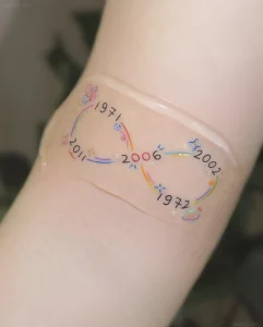 Крупный план татуировки в виде символа бесконечности с датами 1971, 2002, 1972, 2011 и 2006, украшенной радужными линиями и звёздами на коже предплечья
