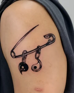 Черно-белая татуировка в стиле реализм: булавка, пронзающая два символа инь-ян, соединённых цепочкой, на плече человека