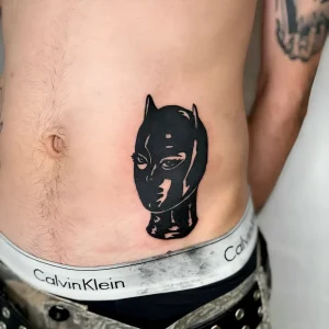 Черно-белая татуировка в стиле blackwork, изображающая маску Бэтвумен, расположенная на правом боку мужчины над поясом Calvin Klein. Работа выполнена в технике плоской заливки с минималистичными деталями.