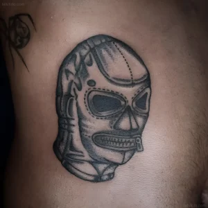 Черно-белая татуировка на коже: маска luchador с молнией по лицу и молчащей губой, символизирующая подавление и контроль в бдсм-контексте